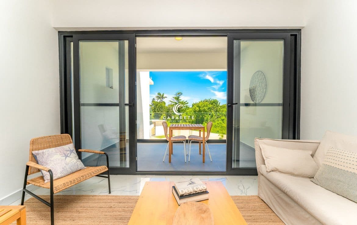 Modern living room with sliding glass doors leading to a balcony with outdoor seating and a view of a tropical garden.